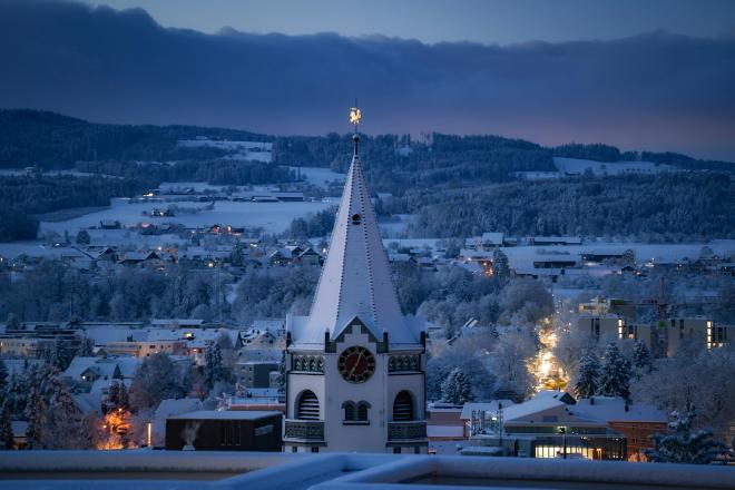A peaceful winter evening settles over a snow-covered European village, where warm Christmas lights glow softly against the blue tones of dusk. In the foreground, a historic church tower rises above the rooftops, crowned with snow and a golden weather vane, evoking a timeless holiday spirit. The surrounding hills and forests are blanketed in white, creating a serene alpine Christmas scene full of calm, warmth, and seasonal magic.
